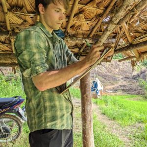 Graduate student in green flannel shirt and pants take notes on small pad in front of thatched roof outdoors