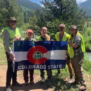 Group of students in green field vests holding a CSU Colorado flag, forest in background