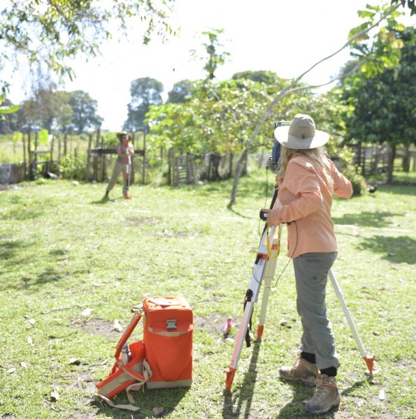 Woman in field clothes in foreground standing next to surveyor equipment standing across green field from another field researcher. Trees in background.