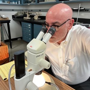 Man looking into stereo microscope at a lab desk.