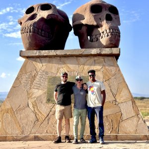 Three men standing in front of monument with two large sculptures of primate skulls found at site.