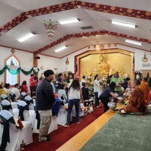 People in Buddhist temple standing to bring offerings to seated monks.