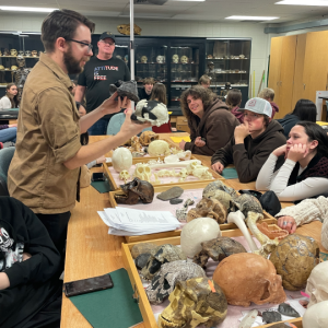 Instructor standing in front of table with skeleton casts with high school students seated.