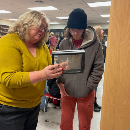 Female professor stands next to high school student looking at glass pressing of small primate skeleton