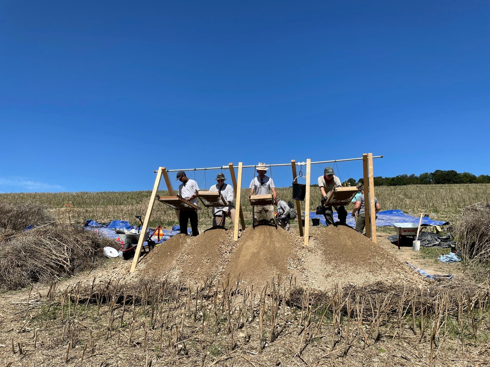 four people sifting materials on small hill of dirt in field