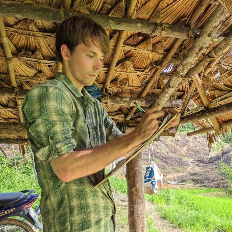 Graduate student in green flannel shirt and pants take notes on small pad in front of thatched roof outdoors