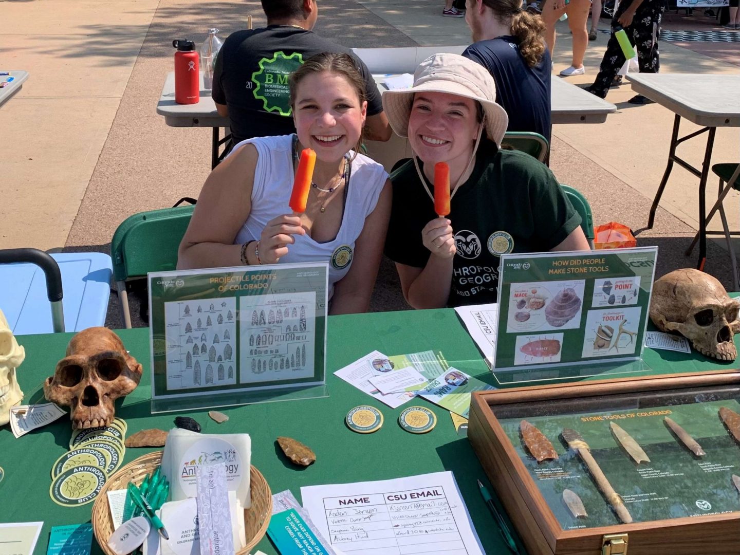 Two students seated at a table with anthropology exhibits and holding popsicles