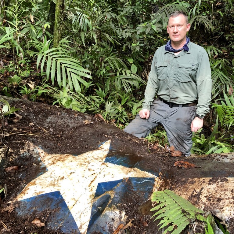 Image of man in long sleeved field shirt and pants standing in forest in Palau in front of wreckage of a fighter plane with white star visible on wreckage