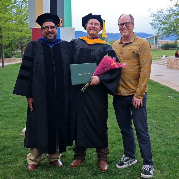 Image show professor, PhD graduate and graduate's father standing outside. Professor and graduate in balck regalia and caps and graduate holds green diploma case